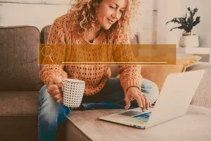 Mulher sorridente usando laptop em casa, representando a indexabilidade do site e sua importância para SEO. Ela segura uma caneca e pesquisa informações.