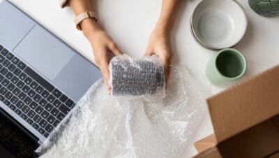 Pessoa embalando produto com papel bolha, ao lado de caixas, copos decorativos e laptop, ilustrando opções de embalagens como filme PVC, stretch e termoencolhível.